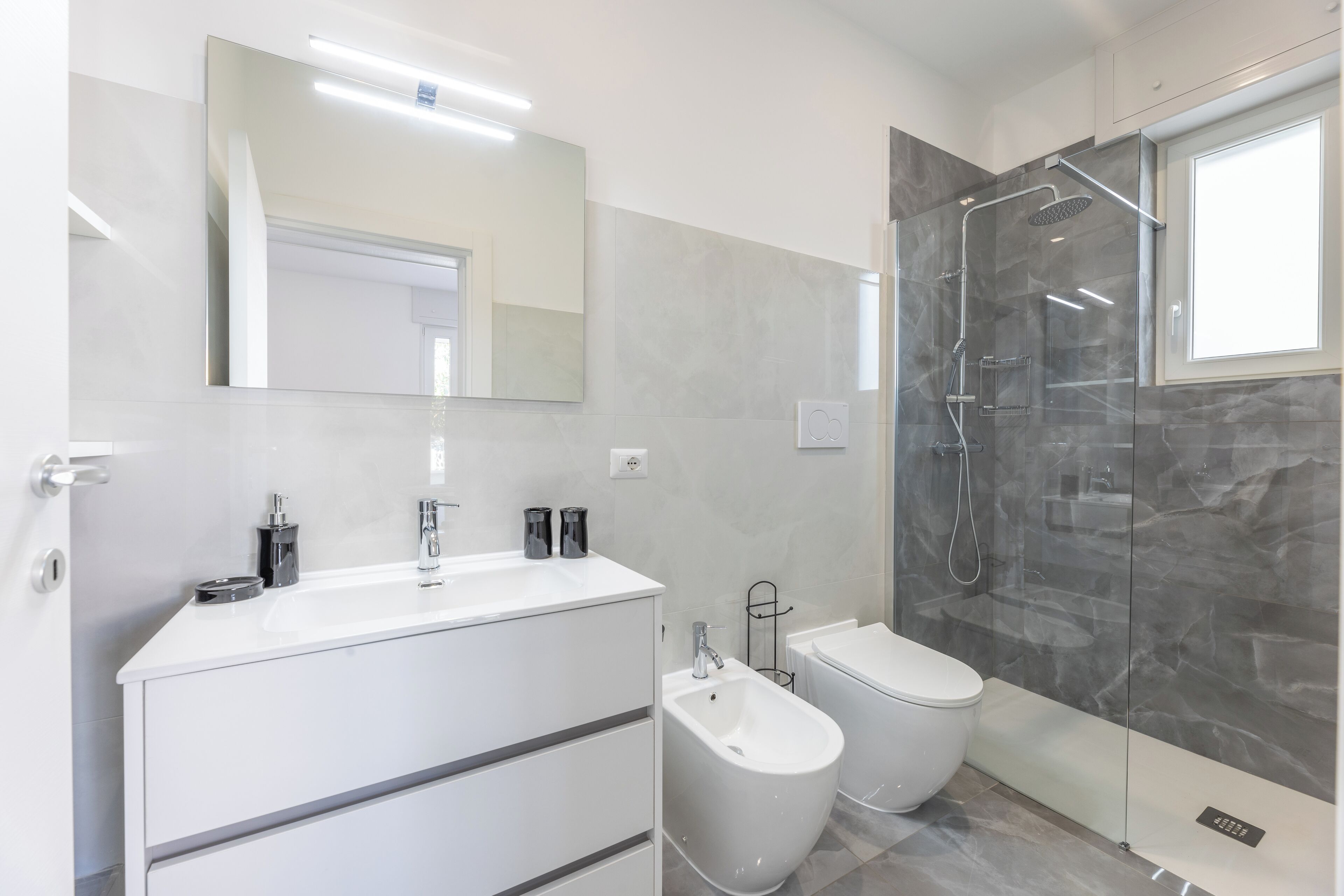 Un baño moderno con tocador blanco, un bidé, un inodoro y zona de ducha con mampara de vidrio.