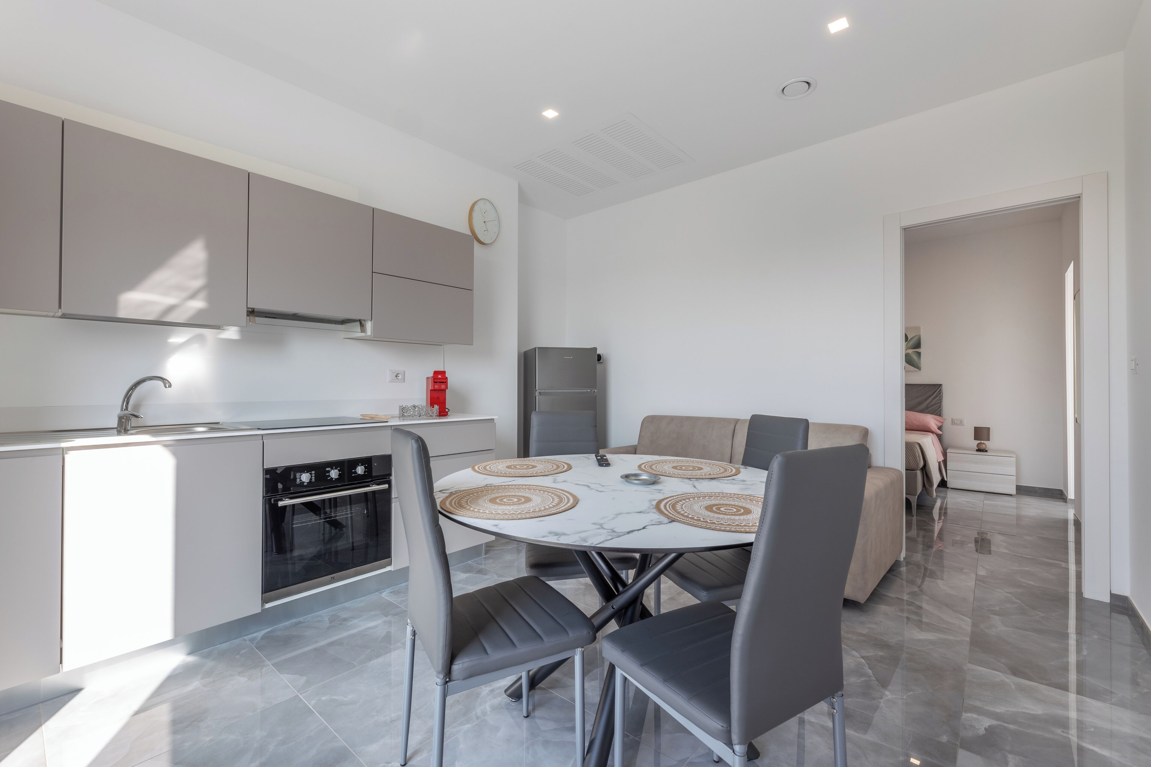 A modern kitchen with grey cabinets, a built-in oven, and a dining area with a marble table and grey chairs.