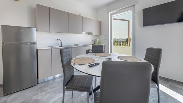 A modern kitchen with grey cabinets, a stainless steel refrigerator, and a dining area with a marble table and grey chairs.