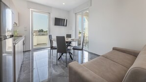 A modern living room with a beige sofa, a dining area with a table and chairs, and a kitchen area with a refrigerator and cabinets.