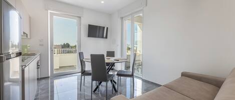 A modern living room with a beige sofa, a dining area with a table and chairs, and a kitchen area with a refrigerator and cabinets.