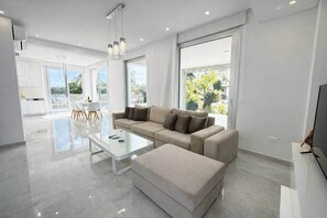 Een moderne woonkamer met een bank, voetenbankje en een glazen salontafel. Er is een eethoek met stoelen en een tafel, en grote ramen die natuurlijk licht bieden.