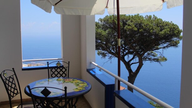 A balcony with a table and chairs, a white umbrella, and a view of the sea and a tree. - From Grandfather Aurelius (Campania)