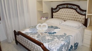 A neatly made bed with a floral bedspread, a wooden headboard with intricate carvings, and a white nightstand beside it. - From Grandfather Aurelius (Campania)