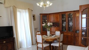 A dining area with a round table, white chairs, and a chandelier. There is a television on a wooden cabinet to the left. - From Grandfather Aurelius (Campania)