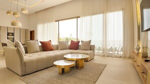 Ein modernes Wohnzimmer mit einer Eckcouch, zwei runden Couchtischen und einem großen Fenster mit leicht transparenten Vorhängen.