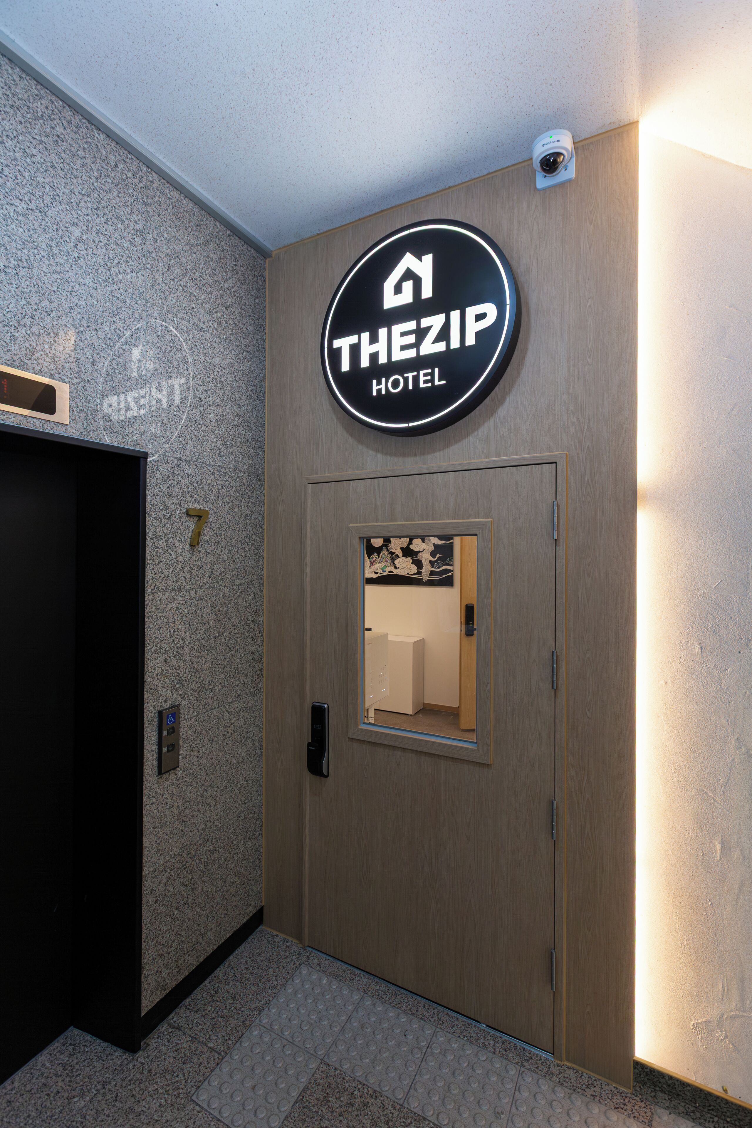 L’entrée d’un hôtel, avec une enseigne indiquant « THEZIP HOTEL », une porte en bois dotée d’une petite fenêtre et une caméra de surveillance placée au-dessus.