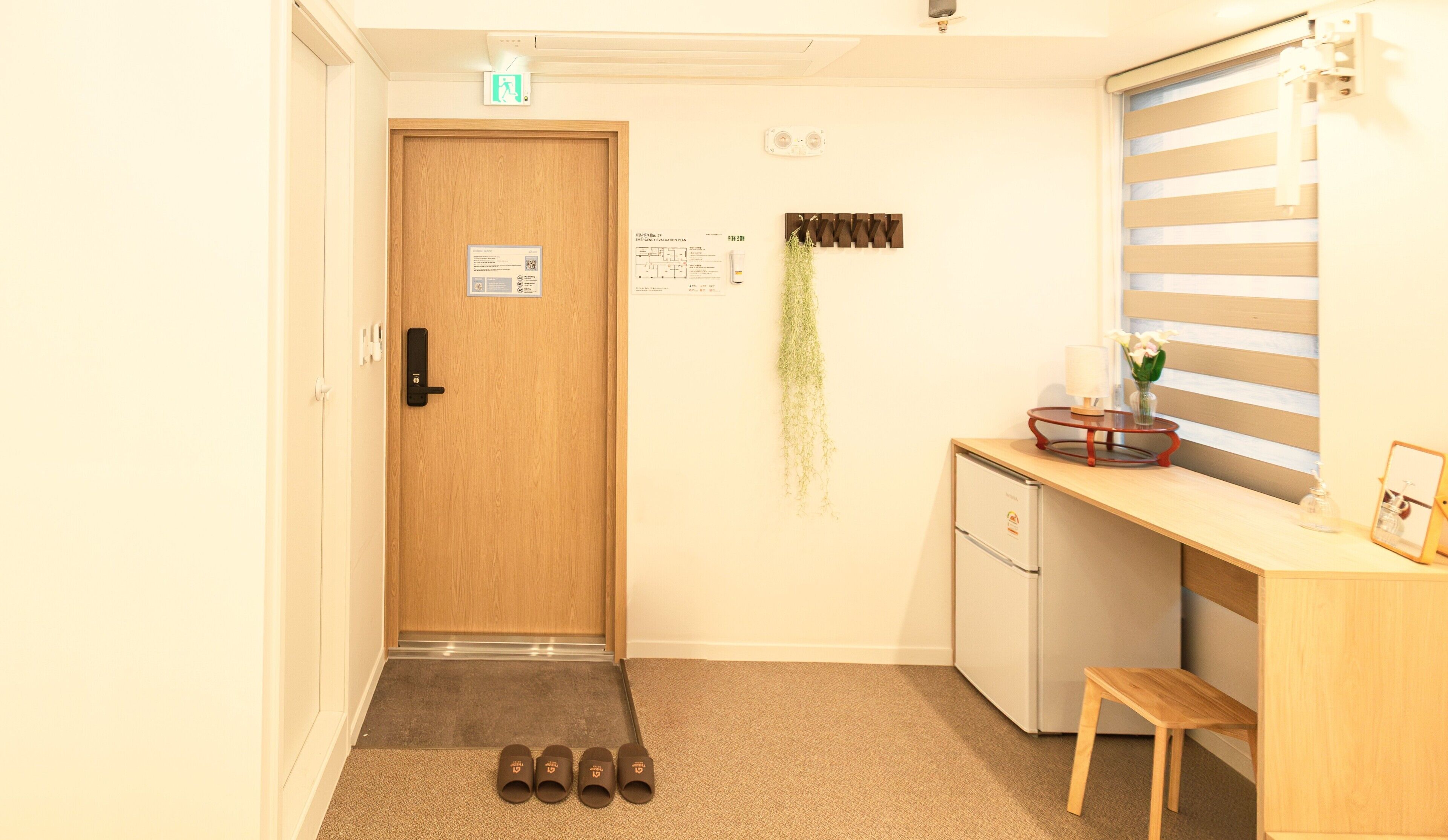 Ein Hotelzimmer mit Holztür, einem kleinen Schreibtisch mit Spiegel, einem Kühlschrank und einer Bank.