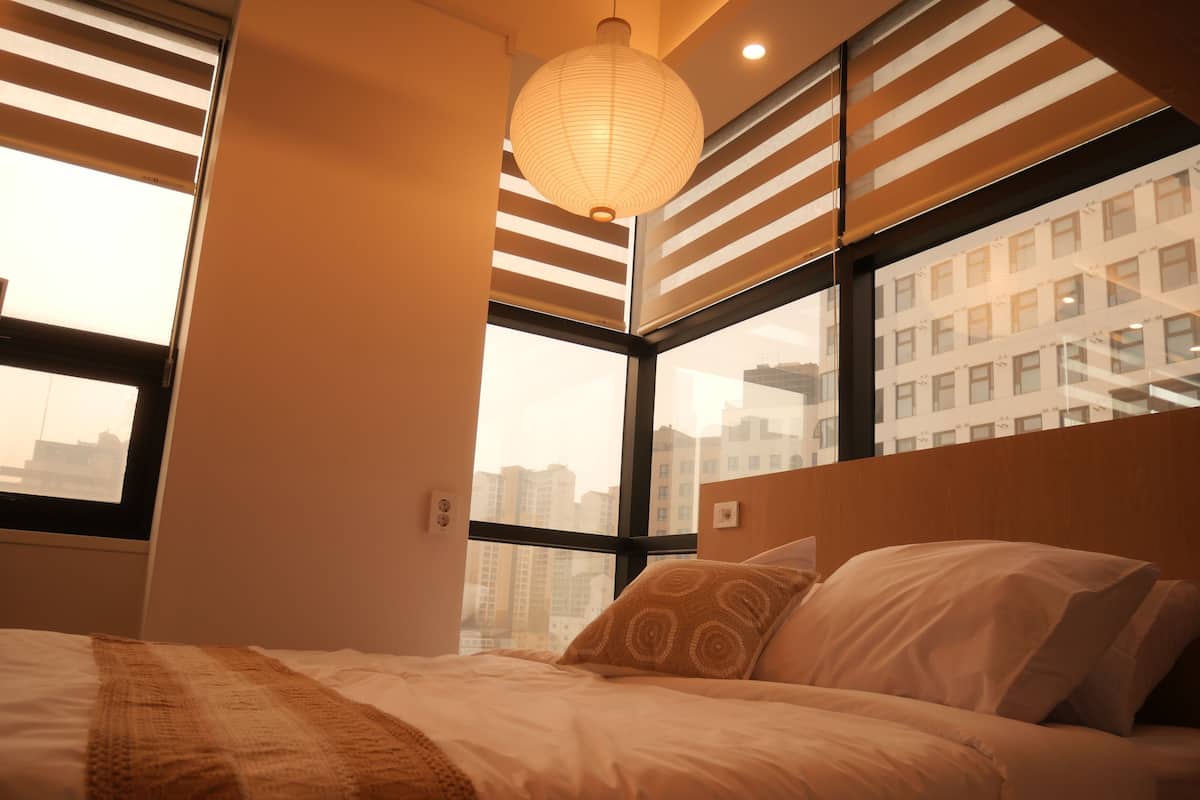 A bedroom with a bed, a hanging light fixture, a window with blinds, and a city view.
