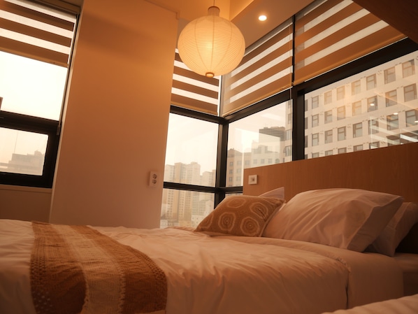 A bedroom with a bed, a hanging light fixture, a window with blinds, and a city view.