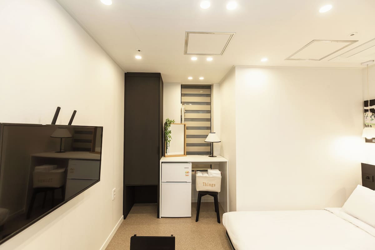 A compact hotel room with a bed, a desk with a computer, a small refrigerator, a chair, and a mirror.
