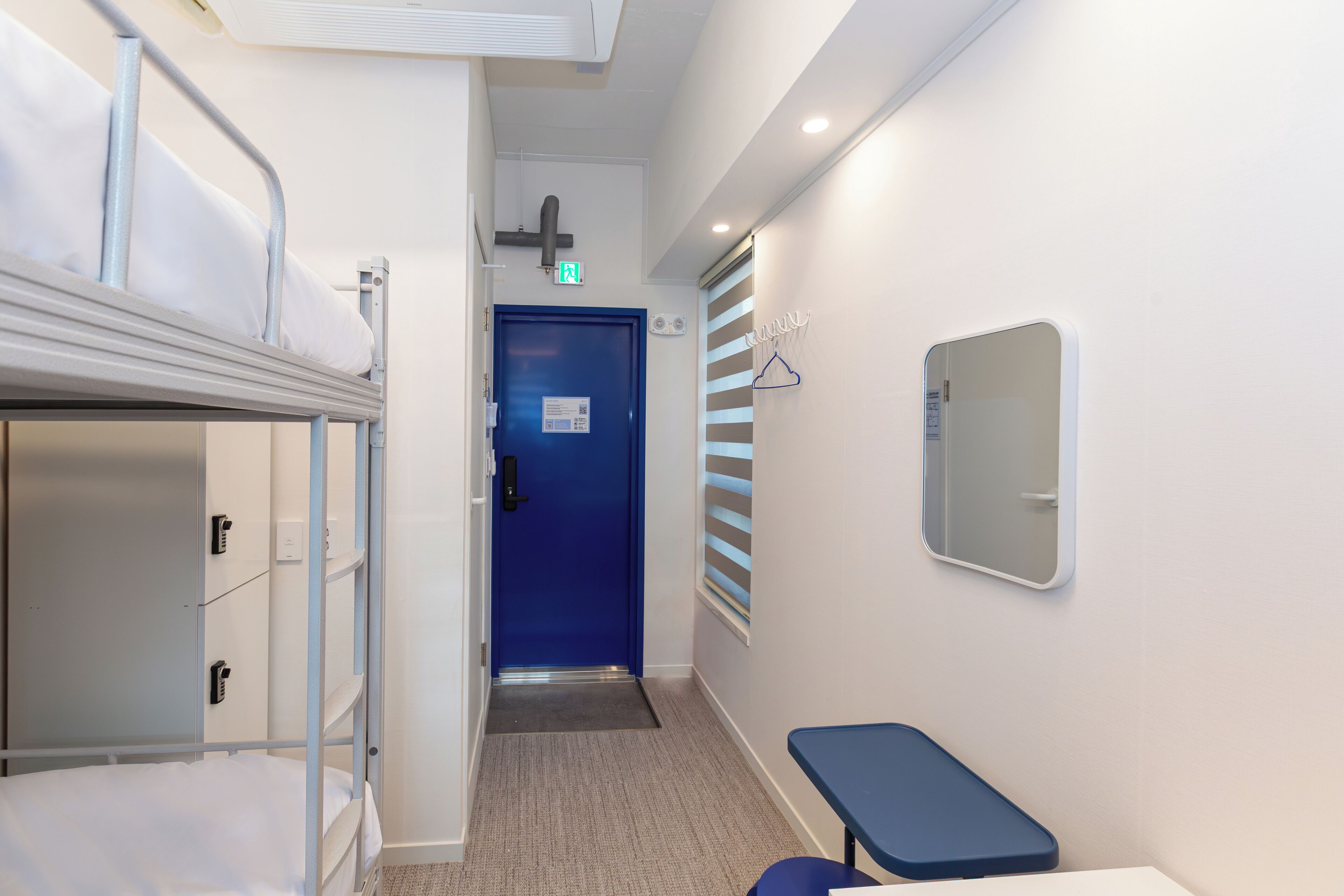 A hotel corridor with a blue door, bunk beds, a small table, and a mirror on the wall.