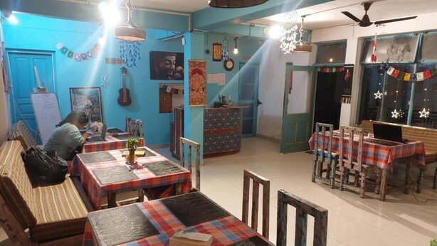 Uma sala de jantar com várias mesas e cadeiras, uma pessoa trabalhando em uma escrivaninha, e diversas decorações nas paredes.