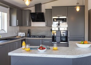 A modern kitchen with grey cabinets, a central island with fruit and drinks, a microwave, and a stove. - 1 bedroom accommodation in Fowey (Fowey)