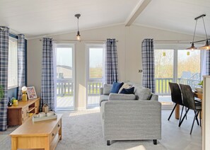 A living room with a sofa, a coffee table, and a dining area with chairs. There are curtains, a ceiling light, and a view through the glass doors. - 1 bedroom accommodation in Fowey (Fowey)