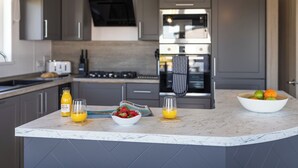 A modern kitchen with grey cabinets, a central island with fruit bowls, and a built-in oven. - Exclusive 3 Bed (Pet) (Fowey)