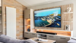A modern living room with a large flat-screen TV mounted on a wooden wall, a sofa, a coffee table, and a bookshelf with a book titled 'SEA & SHORE'. - Exclusive 3 Bed (Pet) (Fowey)