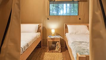 Una tienda de campaña con dos camas de madera, una ventana con vista al bosque, una mesita con una lámpara y un bolso en el suelo.