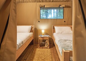A tent with two wooden beds, a window with a forest view, a small table with a lamp, and a bag on the floor. - Safari Tent sleeps 6 (Hayle)