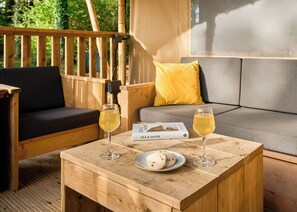 A wooden table with two glasses of white wine, a plate with cookies, and a book titled 'Sea & Shore' on a deck with wooden furniture and a cushion. - Safari Tent sleeps 6 (Hayle)