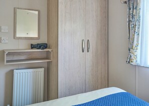 A bedroom with a bed, a wardrobe, a radiator, a shelf with a camera, and a window with curtains. - Standard 3 Bedroom sleeps 8 (Hayle)
