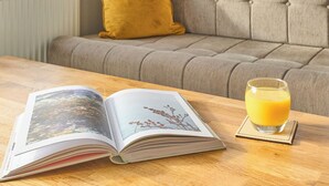Un livre ouvert sur une table en bois, un verre de jus d’orange et un canapé gris avec un coussin de couleur moutarde.