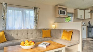 Un salon comprenant un canapé, une table basse avec un panier de fruits, un téléviseur posé sur une étagère en bois, et un coin cuisine avec des placards et des appareils électroménagers.
