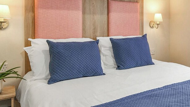 Quarto de hotel com cabeceira de madeira, duas almofadas brancas e duas almofadas azuis. Há duas luminárias de cabeceira e um vaso de planta na mesa de apoio.