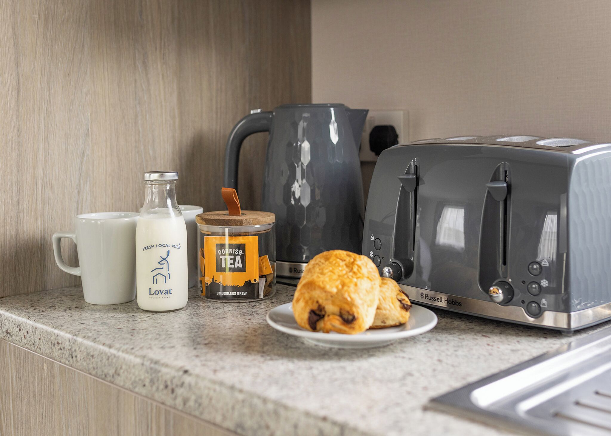 A kitchen counter with a toaster, a mug, a bottle of milk, a jar of tea, and a plate with a pastry.