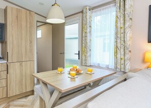 Ein modernes Zimmer mit einem Holztisch, gedeckt für das Frühstück, einem Bett, einem Kleiderschrank und einem Fenster mit Vorhängen.