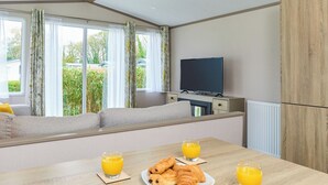 Une chambre moderne avec un canapé près d’une fenêtre, un téléviseur à écran plat, une table à manger avec des croissants et du jus d’orange, et un radiateur.