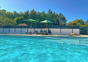 A swimming pool with clear blue water, surrounded by a fence, and shaded by two umbrellas.
