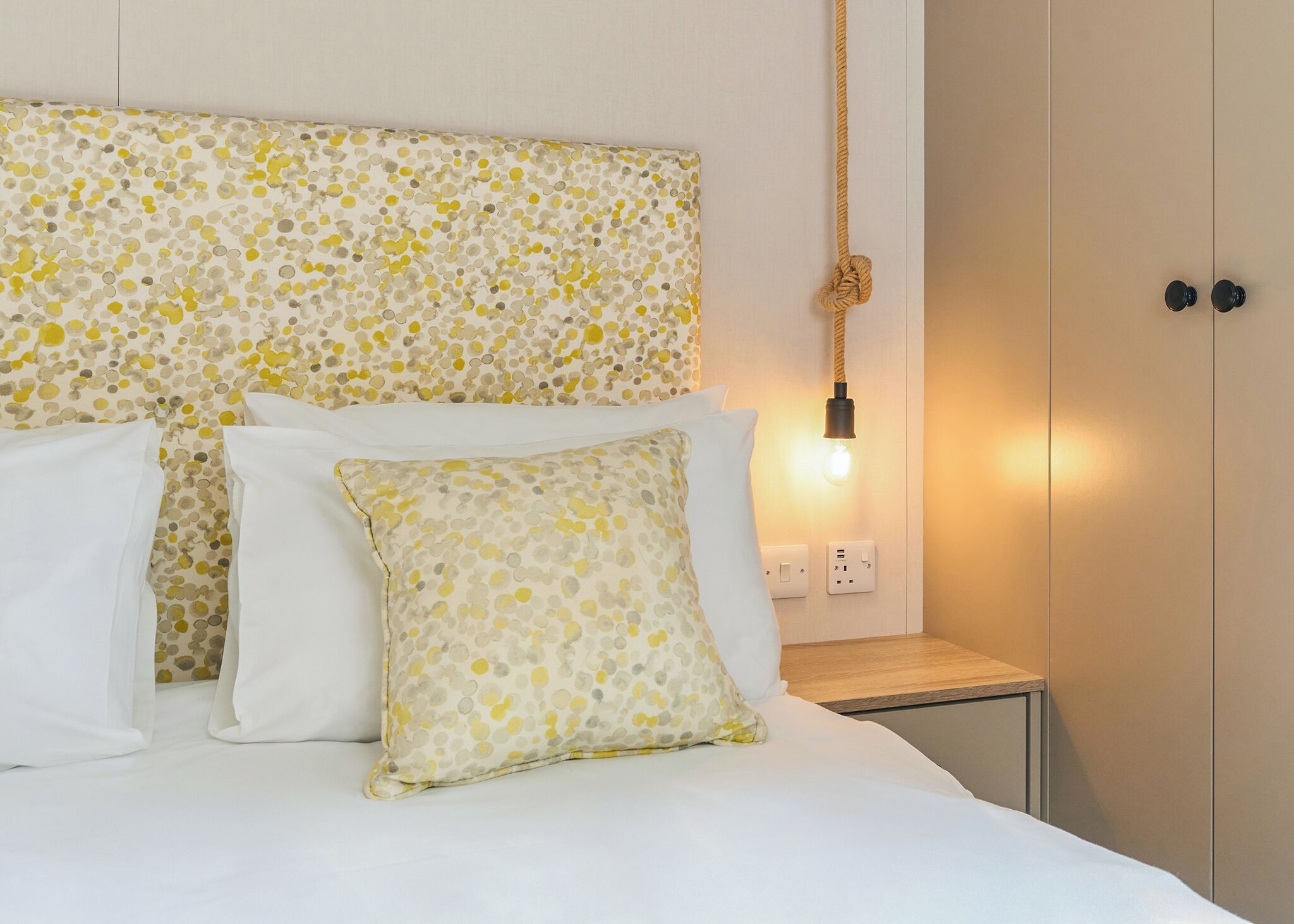 A bed with white bedding and patterned pillows, a bedside table with a lamp, and a wall-mounted light fixture.