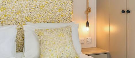 A bed with white bedding and patterned pillows, a bedside table with a lamp, and a wall-mounted light fixture.