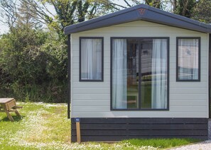 A single-story mobile home with a dark roof, white siding, and black trim. It has two windows with white curtains, a small porch area, and a number sign indicating the unit number.