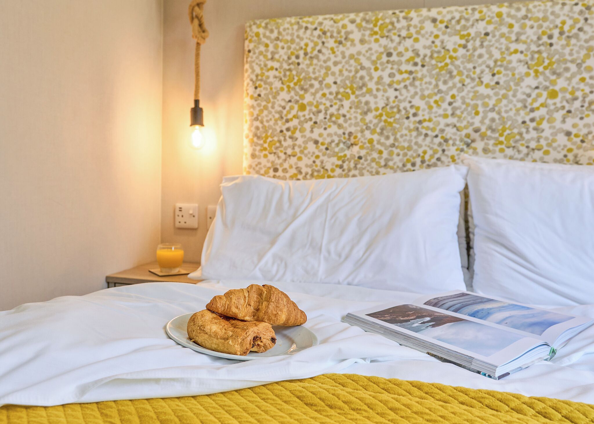 A bed with white linens, two croissants on a plate, a magazine, and a yellow quilt.