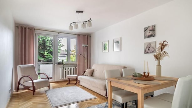 Een moderne woonkamer met een houten eettafel, een bank en een stoel. Er is een groot raam met gordijnen, een radiator en een vloerlamp.