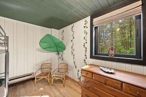 Ein Zimmer mit weißen Wandverkleidungen, Holzboden, zwei Korbstühlen, einer Holzkommode und einem Fenster mit Blick auf einen Wald.