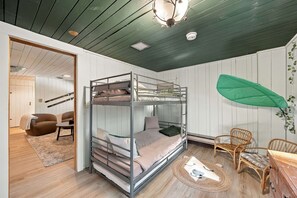 Ein Schlafraum mit Etagenbetten, einem Deckenventilator, einer grünen Blätterdeko und einem Essbereich mit Korbstühlen.