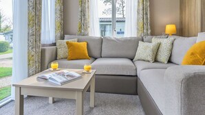 A modern living room with a grey sectional sofa, a small table with a magazine, two glasses of orange juice, and a lamp on a wooden side table. - Superior 3 Bedroom sleeps 8 (Hayle)