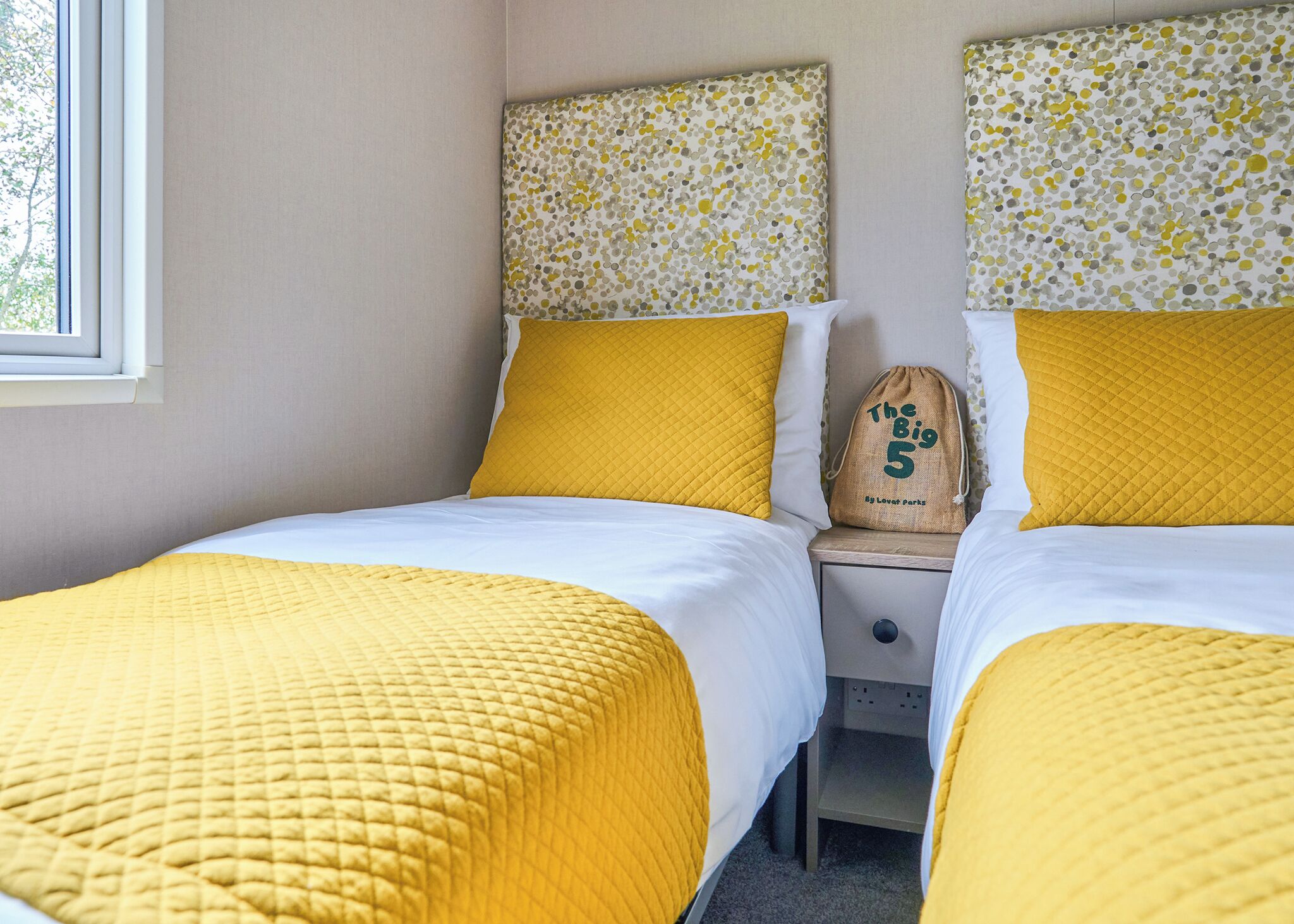 A room with two beds, each with a yellow quilted bedspread and matching pillows, a small bedside table with a lamp, and a window with a view of trees.