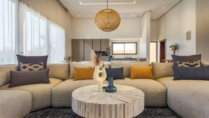 Ein modernes Wohnzimmer mit einem beigefarbenen Ecksofa, gemusterten Kissen, einem runden Couchtisch und einer hängenden Lampenschirm aus Rattan.