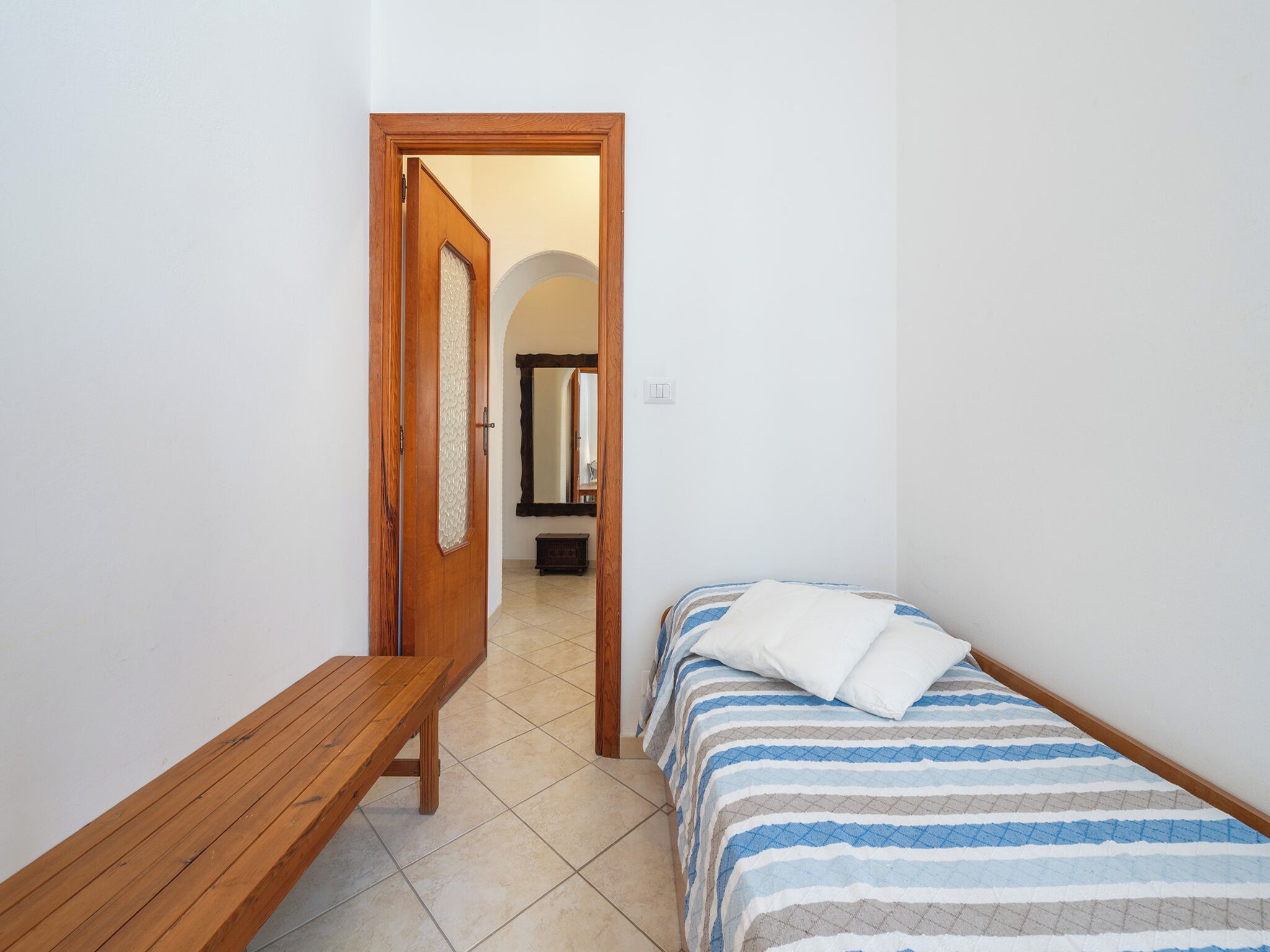 Ein schmaler Flur mit einer hölzernen Bank, einem Bett mit gestreifter Bettwäsche und einem Spiegel an der Wand.