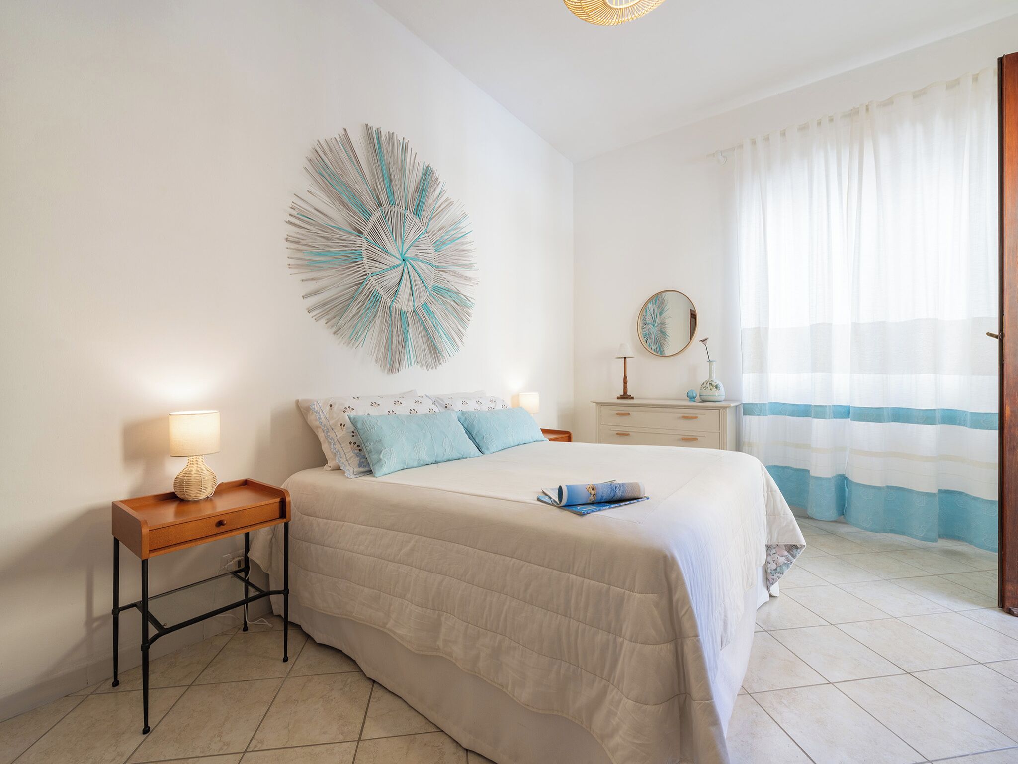 A bedroom with a bed, bedside table, lamp, wall art, dresser, and curtains.