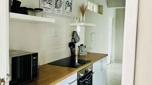 Een moderne keuken met witte kasten, een houten aanrecht, een magnetron en een zwarte spatwand. Er hangen afbeeldingen in lijsten aan de muur en een zwarte deurmat met witte tekst.