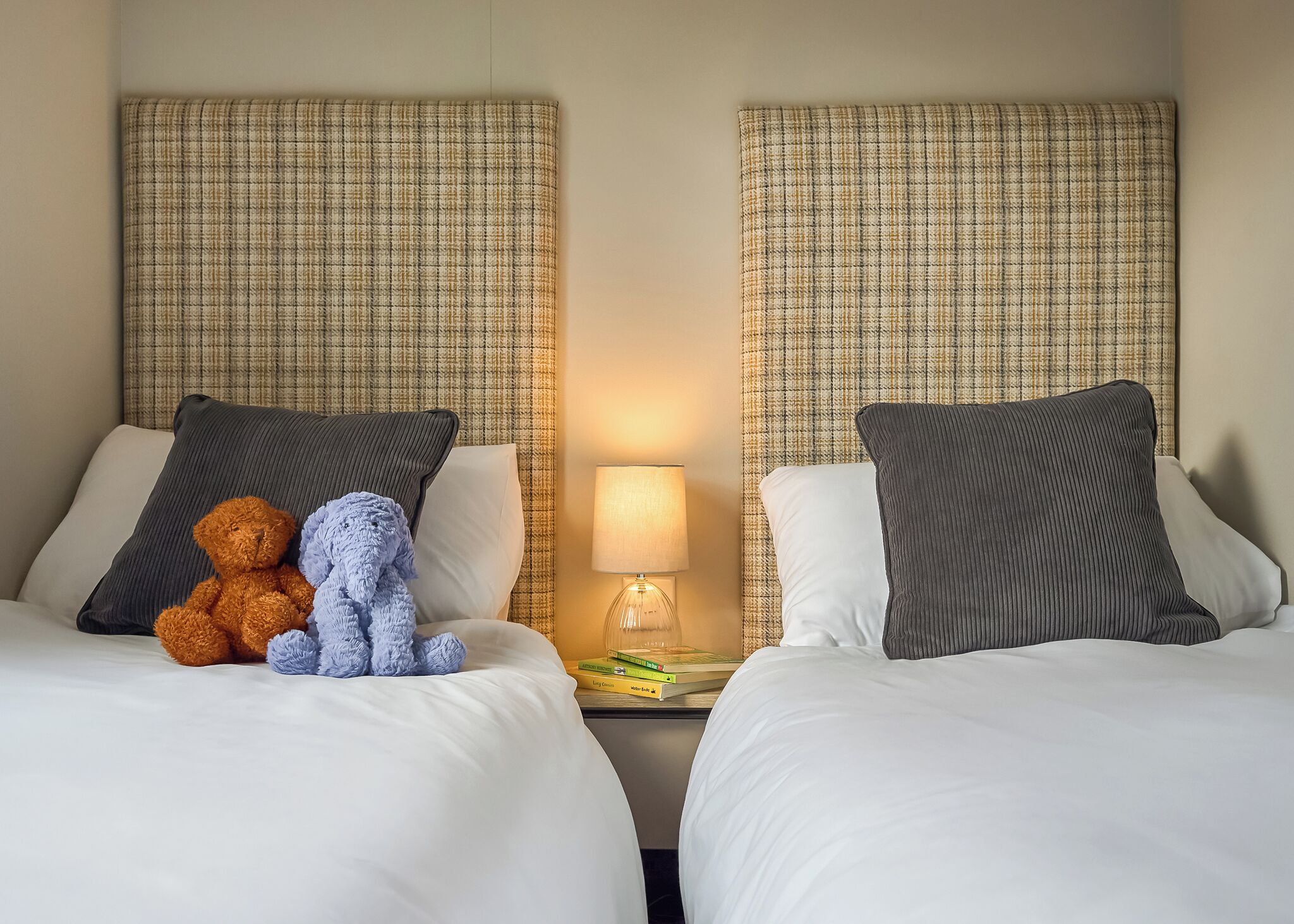 Two beds with headboards, pillows, a teddy bear, a lamp, and a nightstand with books.