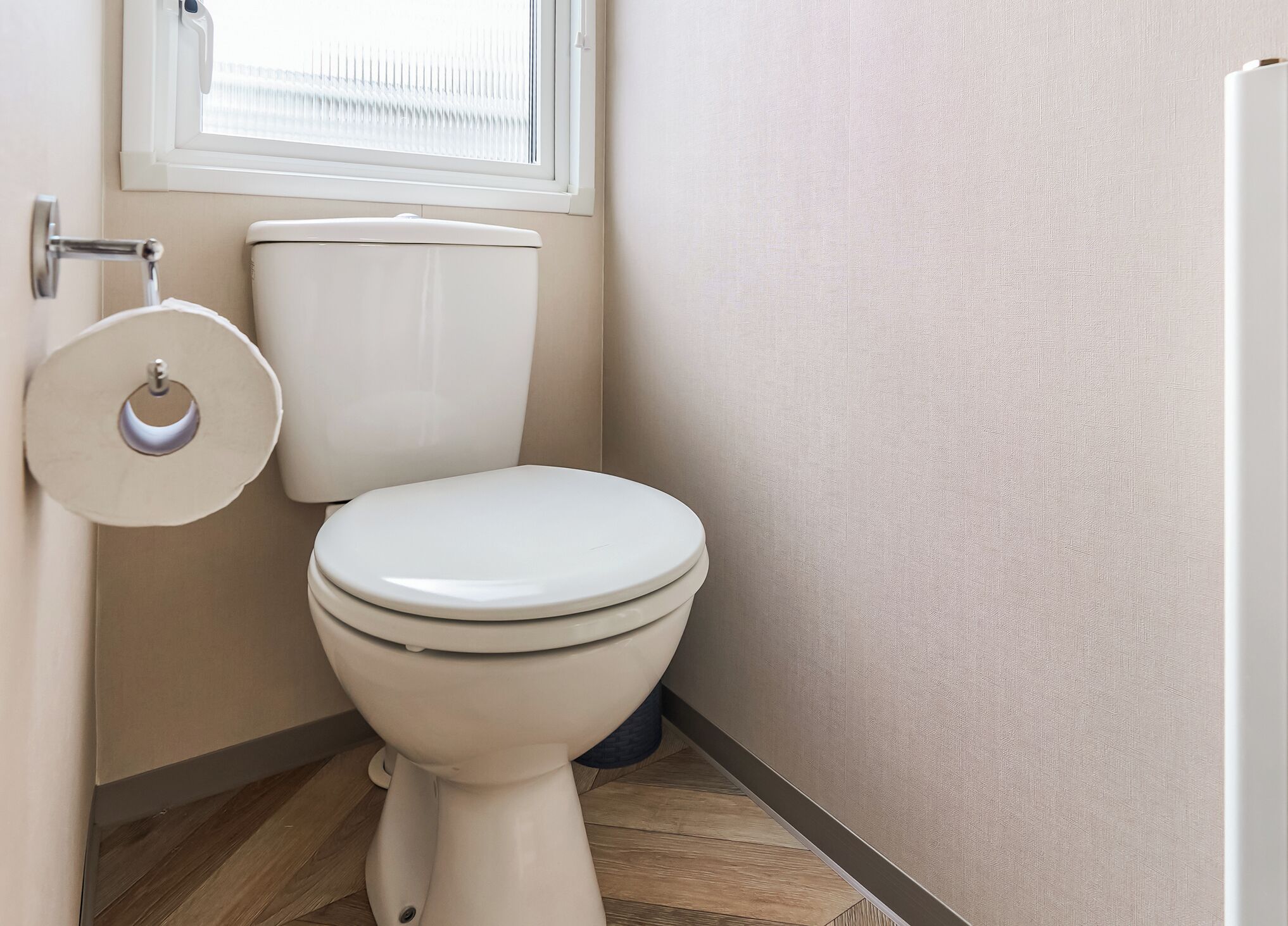 Een toilet met een witte deksel, een toiletrolhouder en een raam erboven.