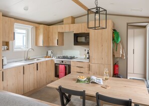 A modern kitchen with wooden cabinets, a gas stove, a microwave, and a dining area with a table and chairs. - Luxury Holiday Home 3 bed (Hayle)
