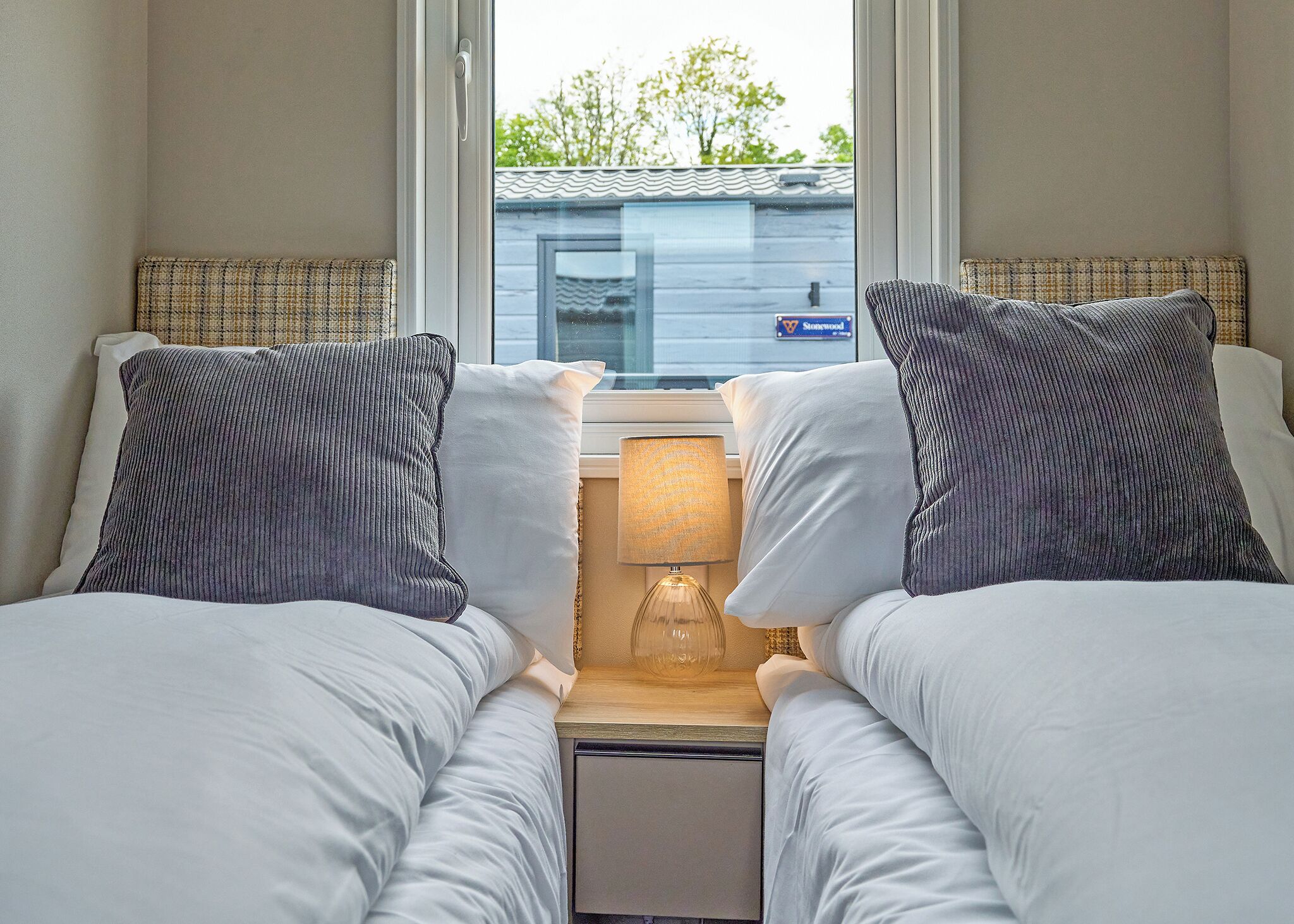 A hotel room with two beds, a nightstand with a lamp, and a window with a view of a building and trees.