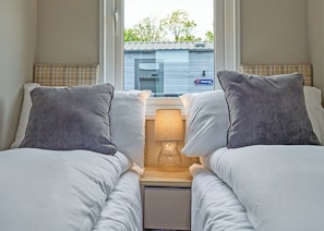 A hotel room with two beds, a nightstand with a lamp, and a window with a view of a building and trees. - Luxury 3 bedroom Hot Tub sleeps 8 (Hayle)